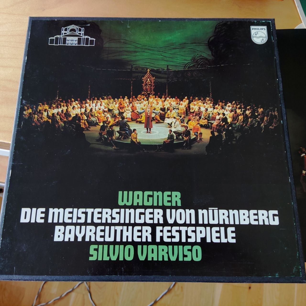 Aufnahme des Covers der 5LP Box von Richard Wagner - Die Meistersinger von Nürnberg - Bayreuther Festspiele 1974 - Dirigent Silvio Varviso. Das Bild zeigt eine Szene der Oper vom Festplatz des Johannisfest bei der die Zusehenden um eine runde Bühne in der Mitte herum sitzen. In der Mitte steht ein Sänger und ihm zu Füßen sitzen außen Musikanten. Im Innenbereich der Bühne sitzen noch drei andere Personen auf Stühlen.